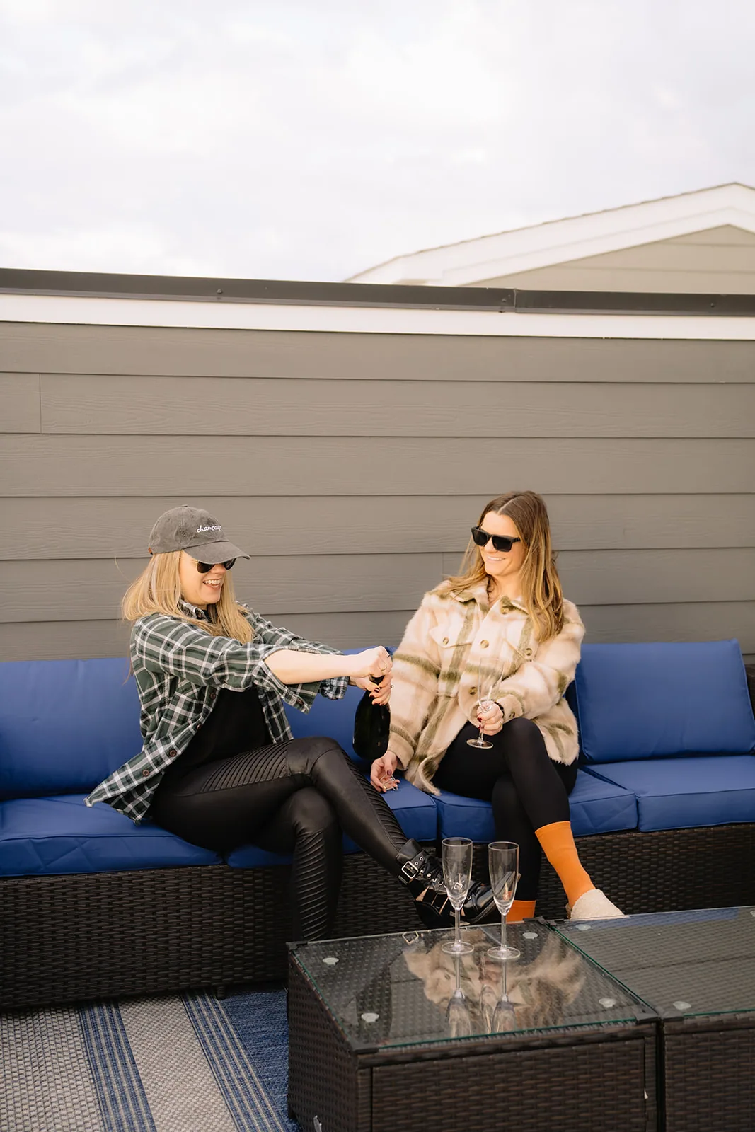Two women in flannel toasting on a blue rooftop sectional with a glass topped table
