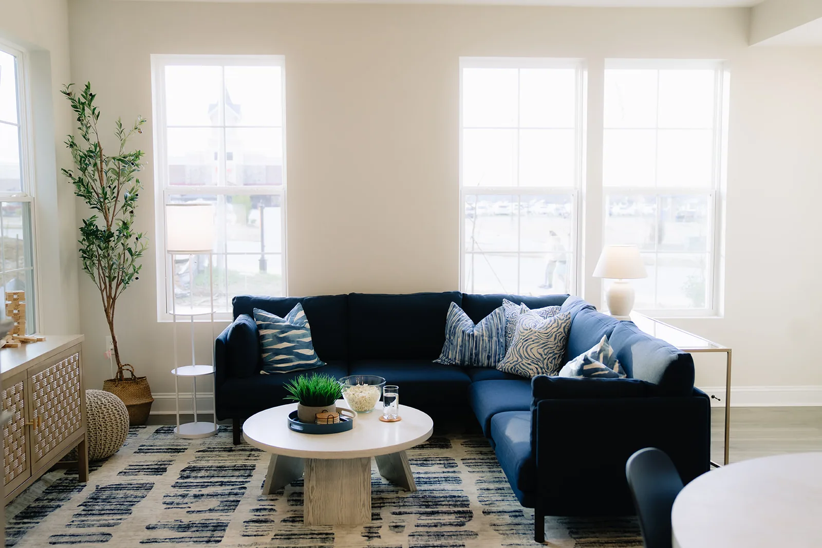 Navy sectional under tall windows with a patterned rug, olive tree and woven console