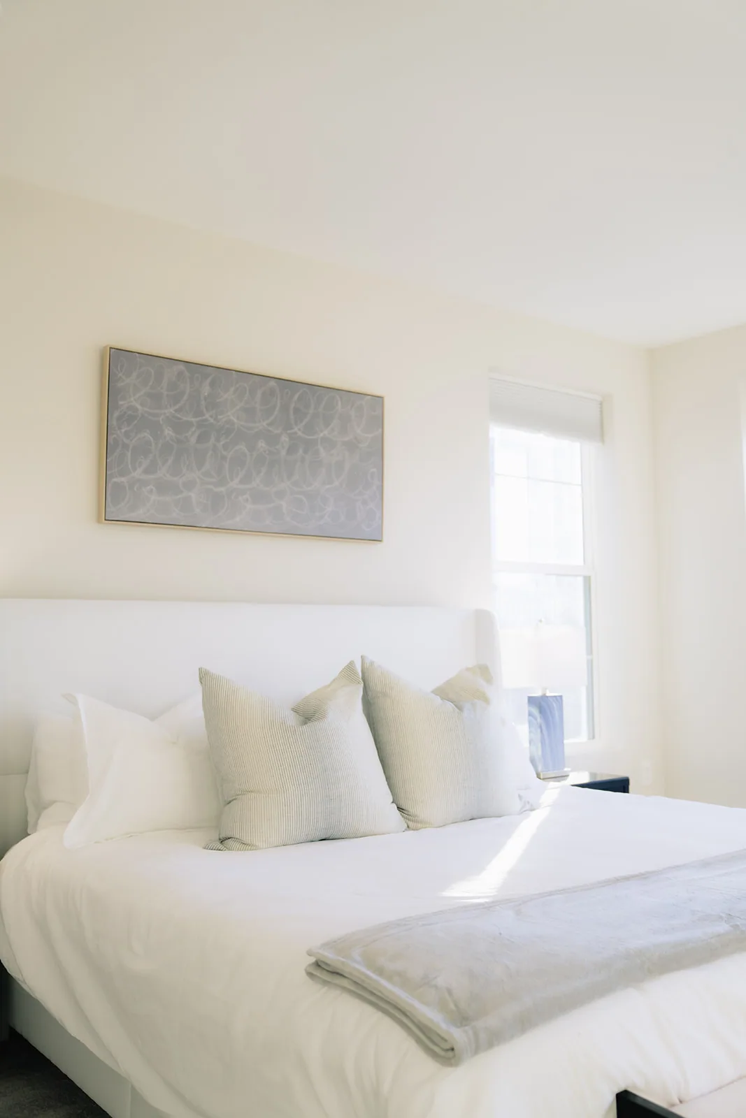 White bed with pale cushions and a folded gray throw at its foot below gray art