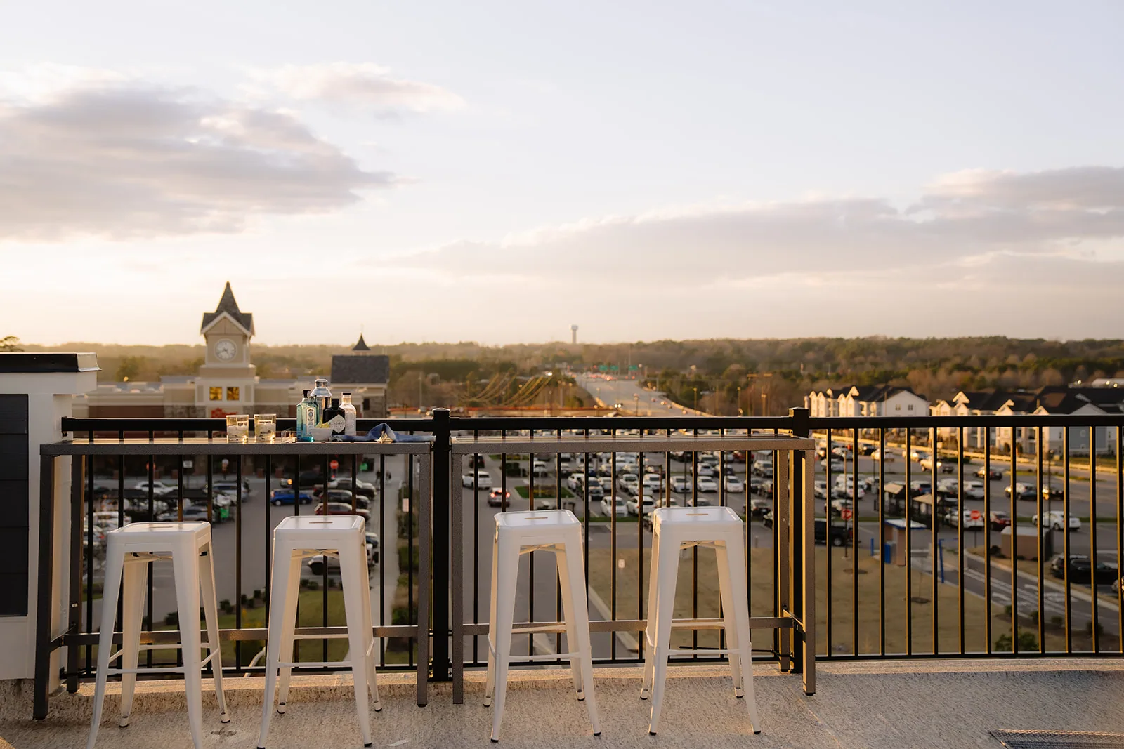 Four white bar stools at a rooftop rail overlooking a lit shopping center at dusk