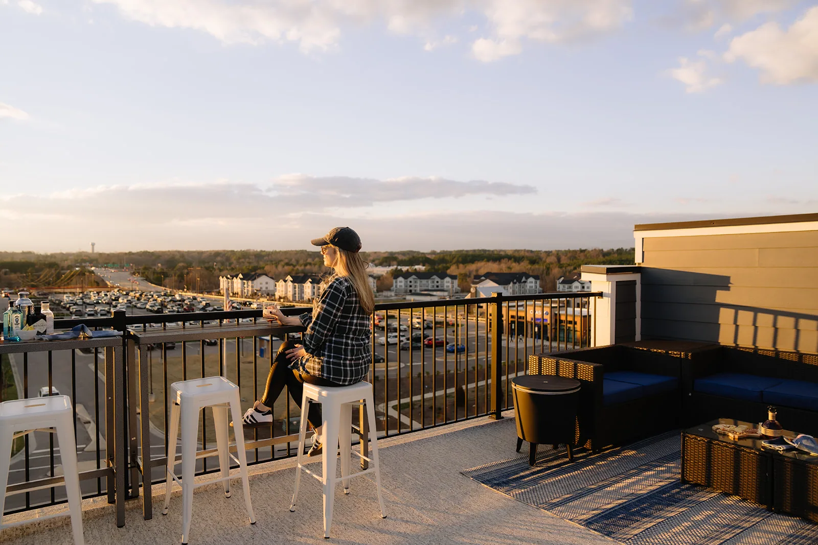 Woman seated on a bar stool at a rooftop rail looking out over a town at dusk