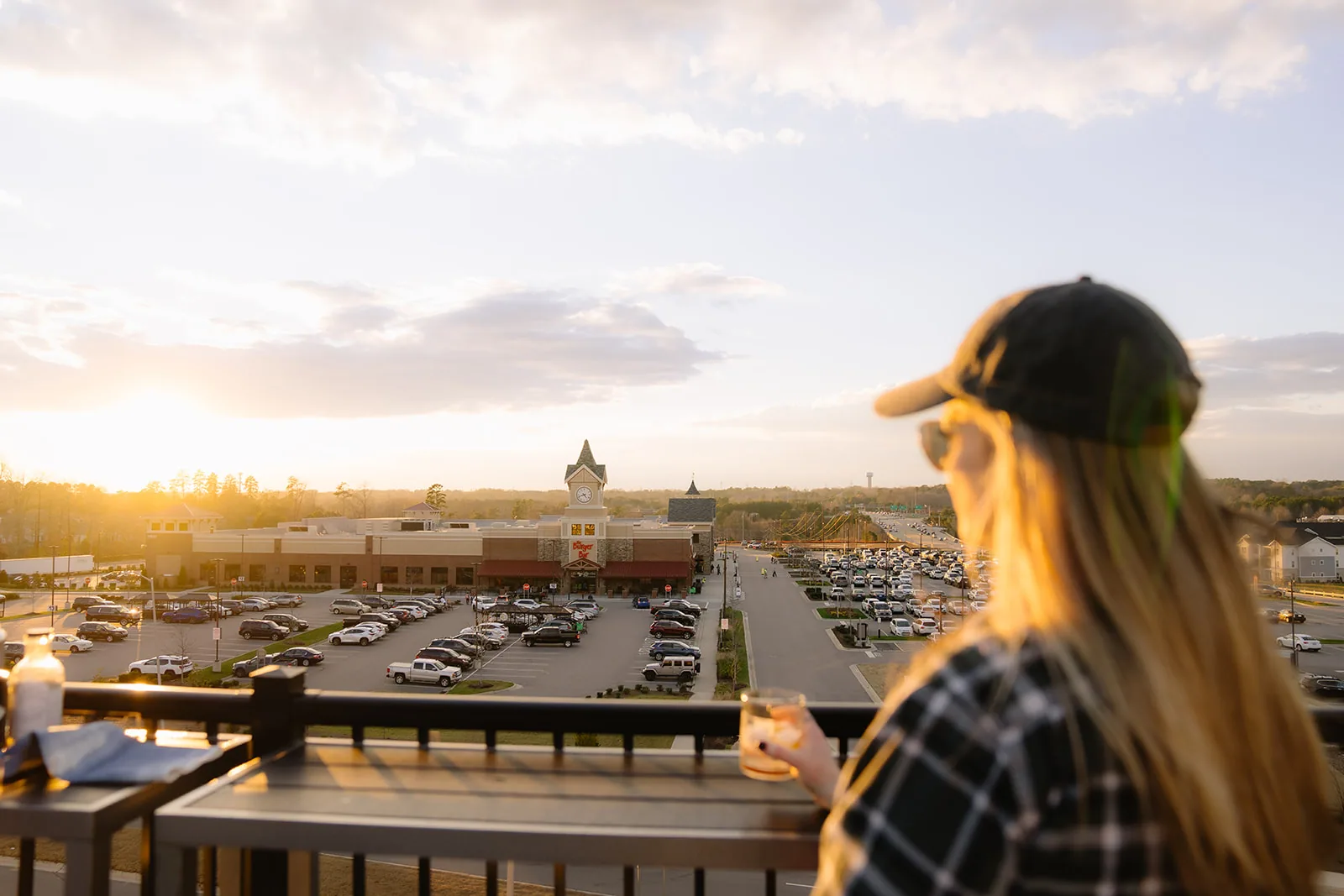 Woman at the rooftop rail with a glass, the sunlit shopping center below