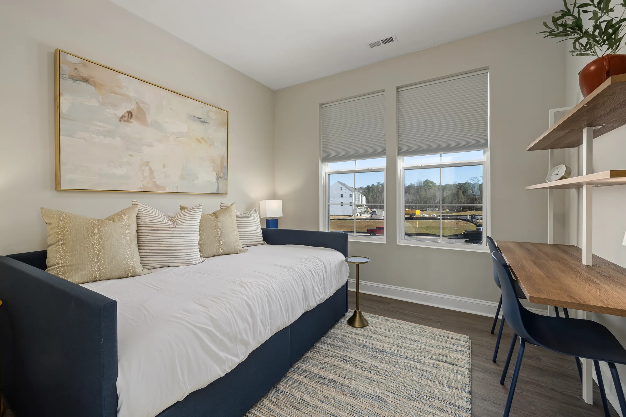 Navy daybed with white bedding under a framed abstract, with a desk and chairs at the window