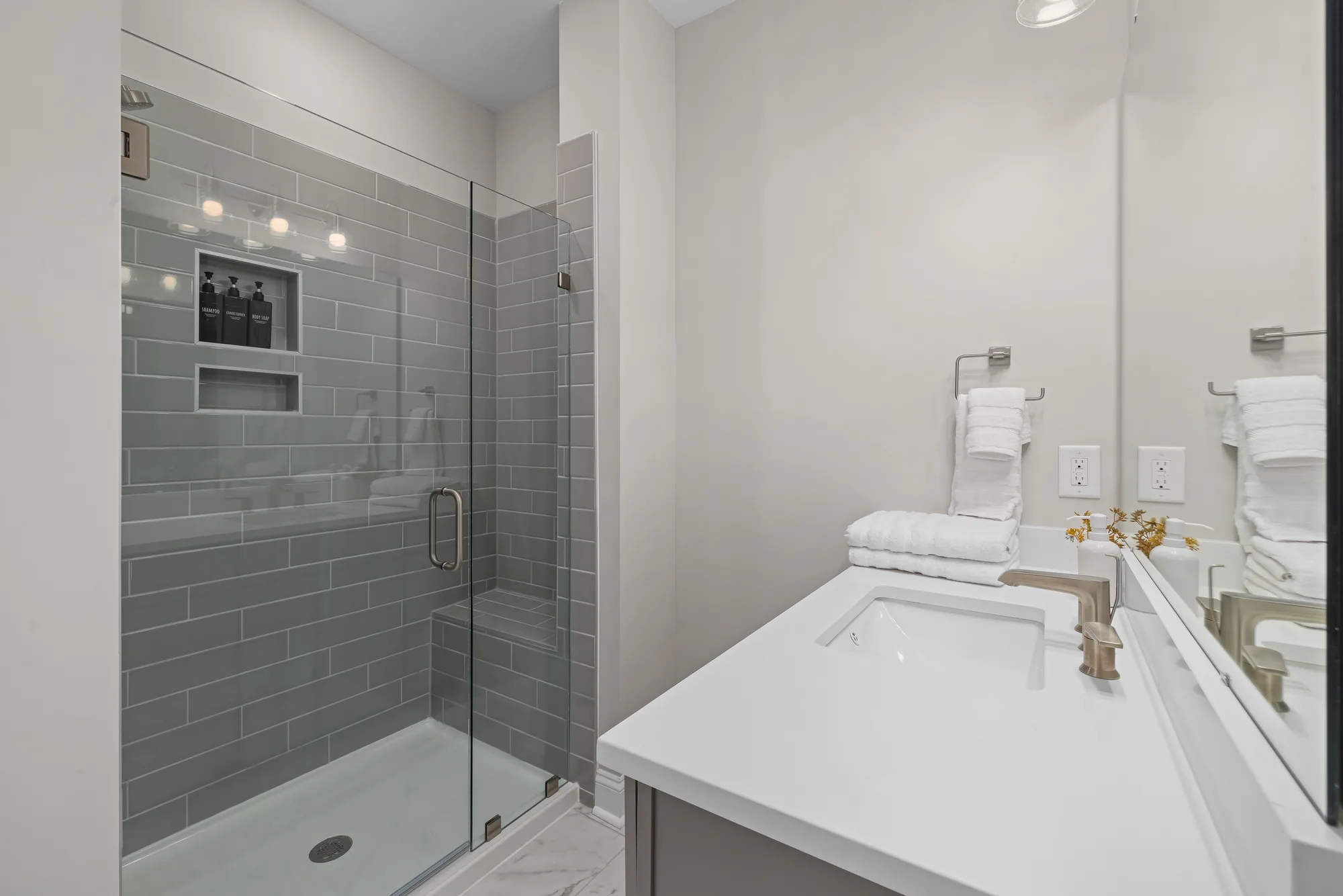 Walk in glass shower with gray subway tile and a niche, beside a white vanity