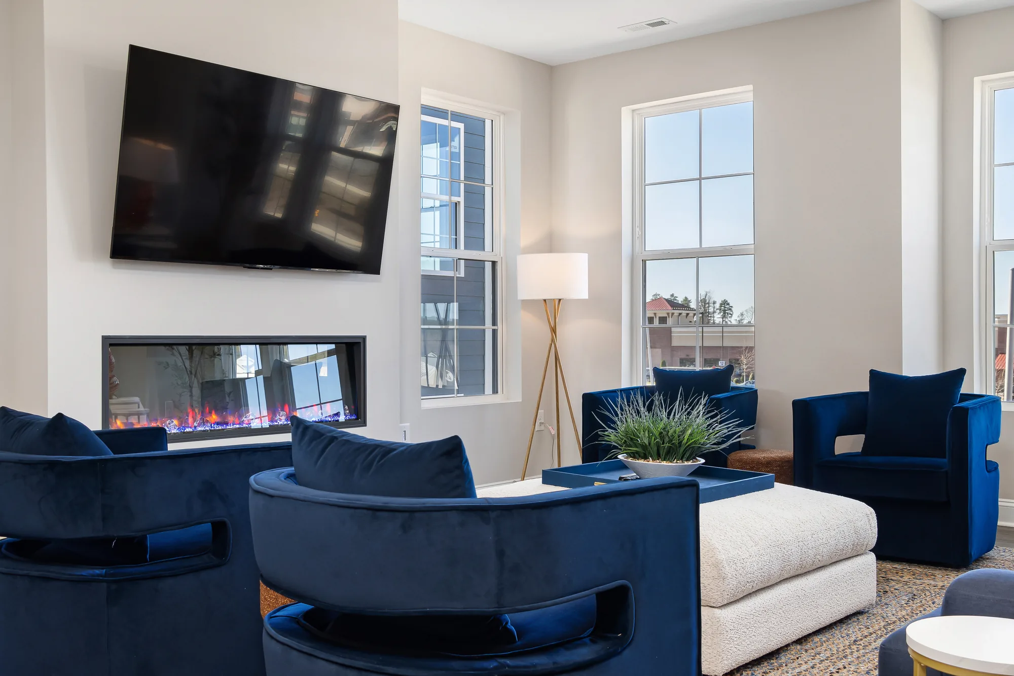 Television above a linear fireplace with blue velvet chairs and a white ottoman in front