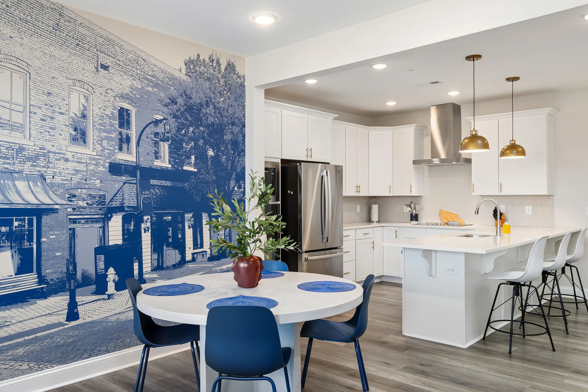 Round white dining table with blue chairs in front of the blue Wake Forest mural, kitchen beyond