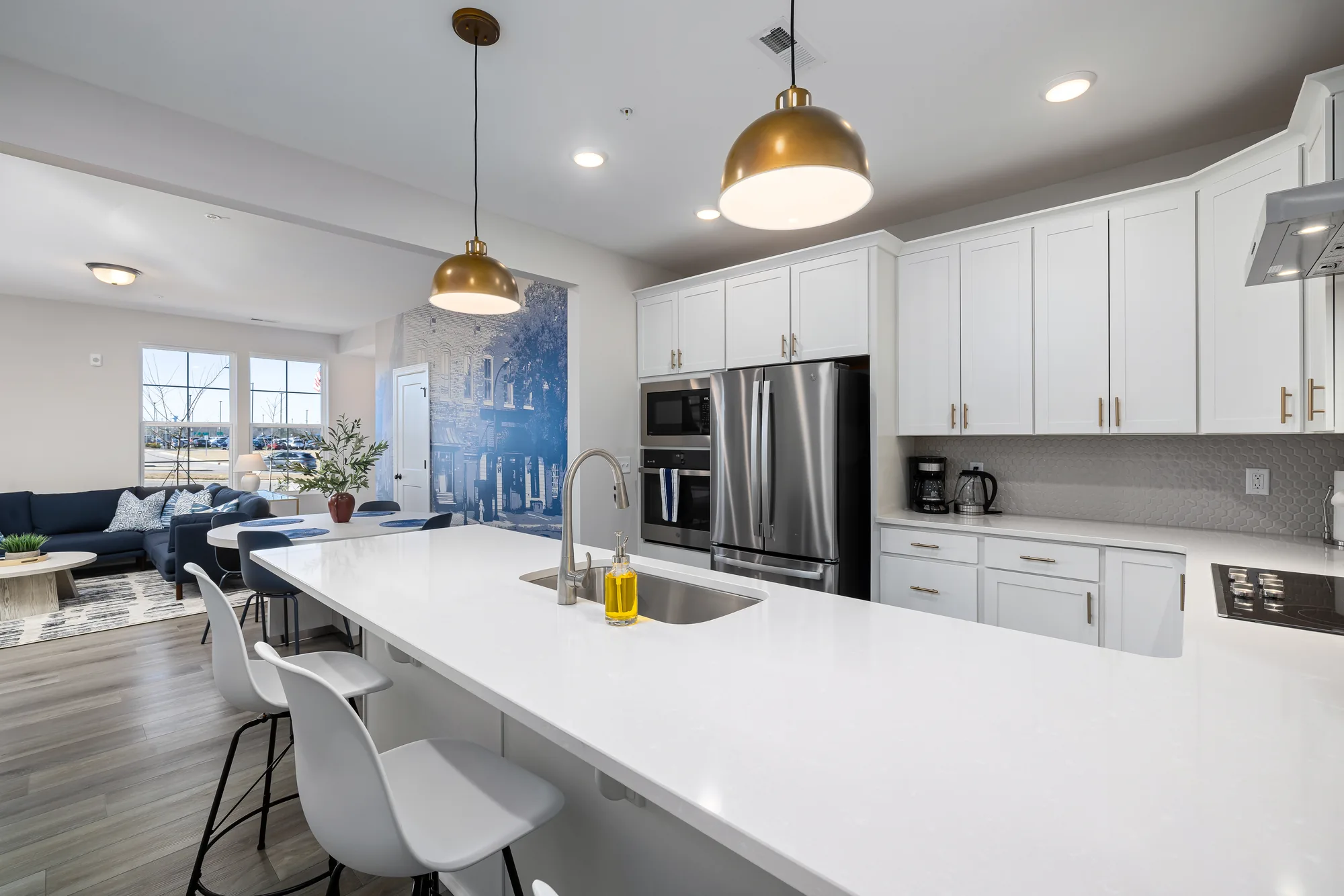 Wide white kitchen with gold pendants and a long island, the mural and living area beyond